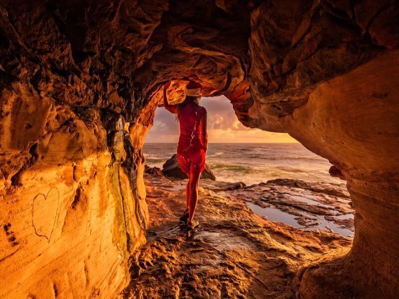 A woman in a red dress is standing in a cave overlooking the ocean.
