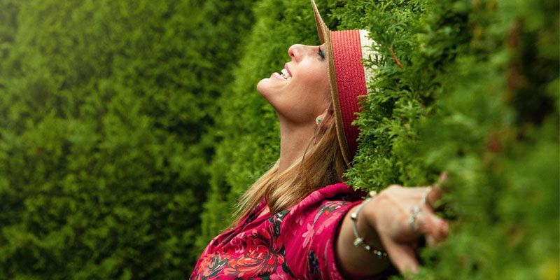 A woman is standing in front of a hedge with her arms outstretched.
