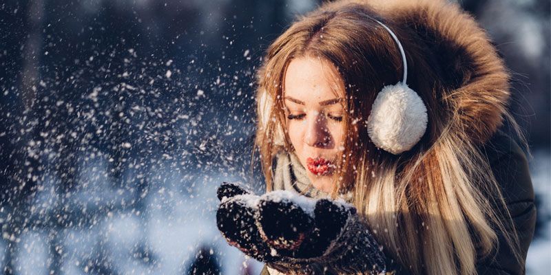 A woman wearing ear muffs is blowing snow into the air.