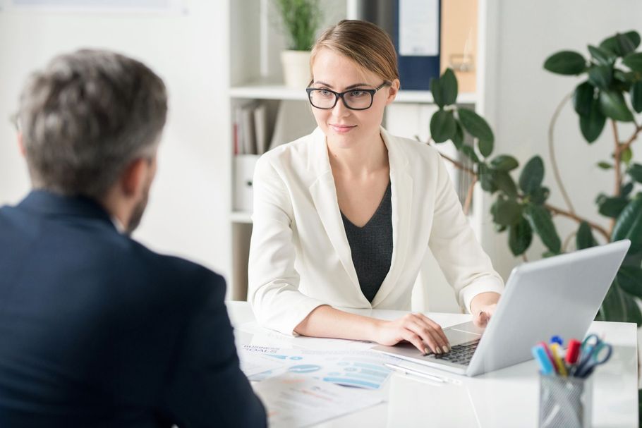Una donna con gli occhiali parla con un uomo dall’altra parte di una scrivania, in un ufficio luminoso, con un portatile aperto lì accanto