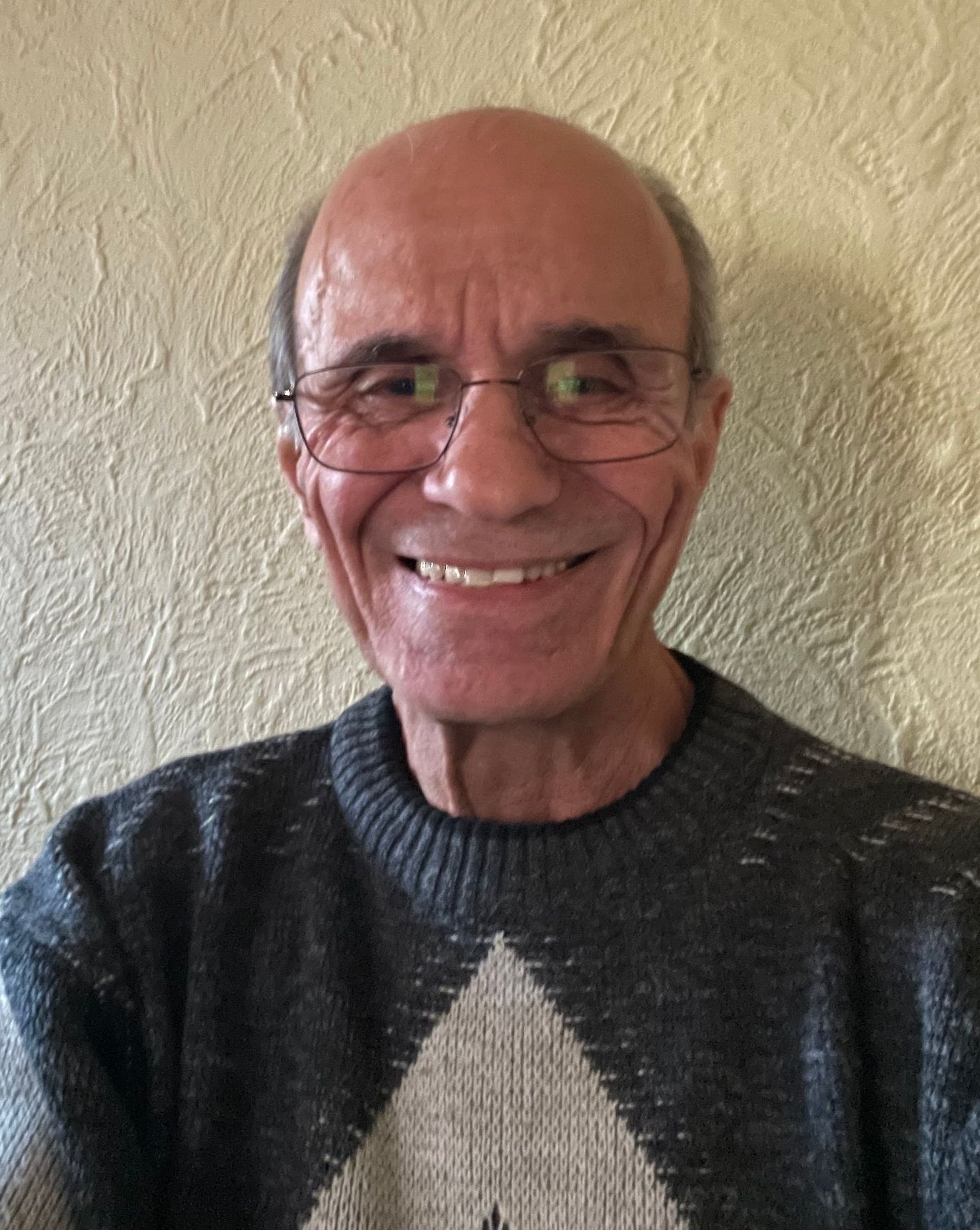 Smiling man with glasses, wearing gray sweater, against a textured wall.