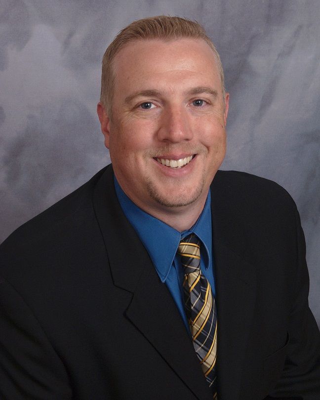 Man in a suit smiles at the camera, with a blue shirt and patterned tie.