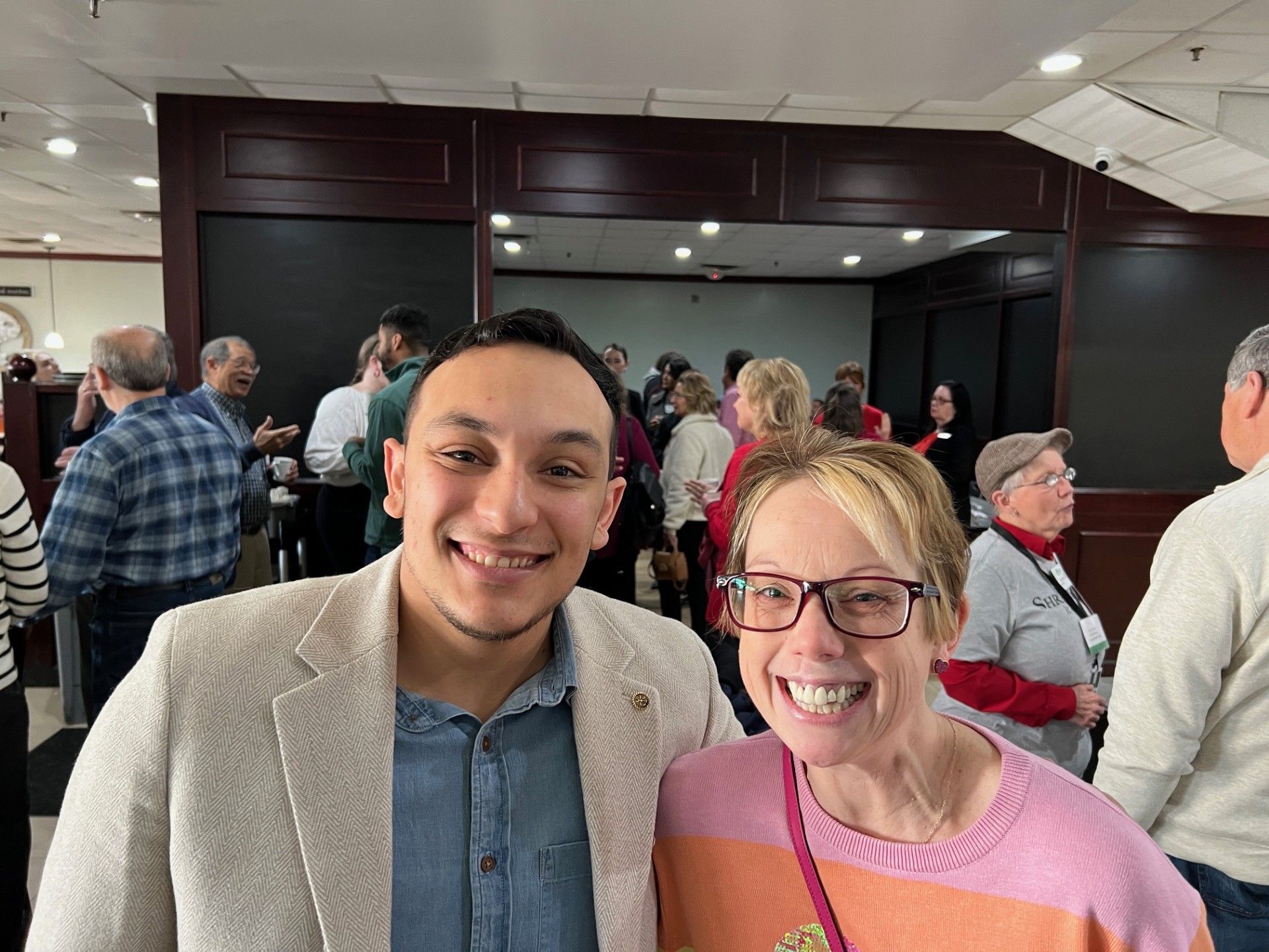 Two people smiling for a photo, other people in background. Indoors, likely event.