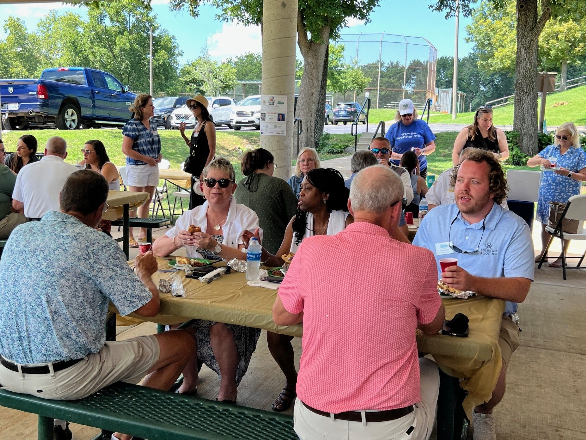 People seated at picnic tables eating outdoors under a pavilion. Blue truck and baseball field visible.