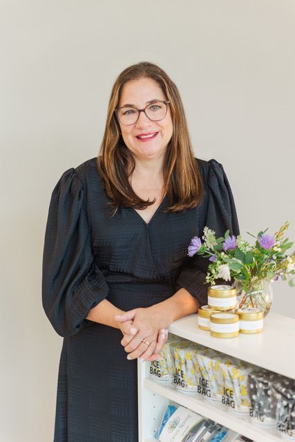 Woman in black dress with glasses, smiles near a table with products and flowers.