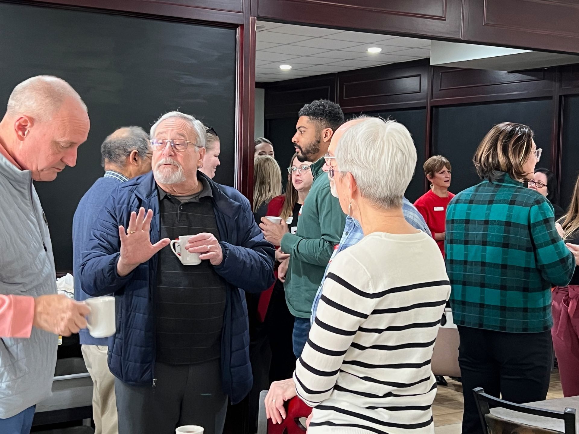 People in a room, some holding mugs, engaged in conversation. Indoor setting with dark walls.