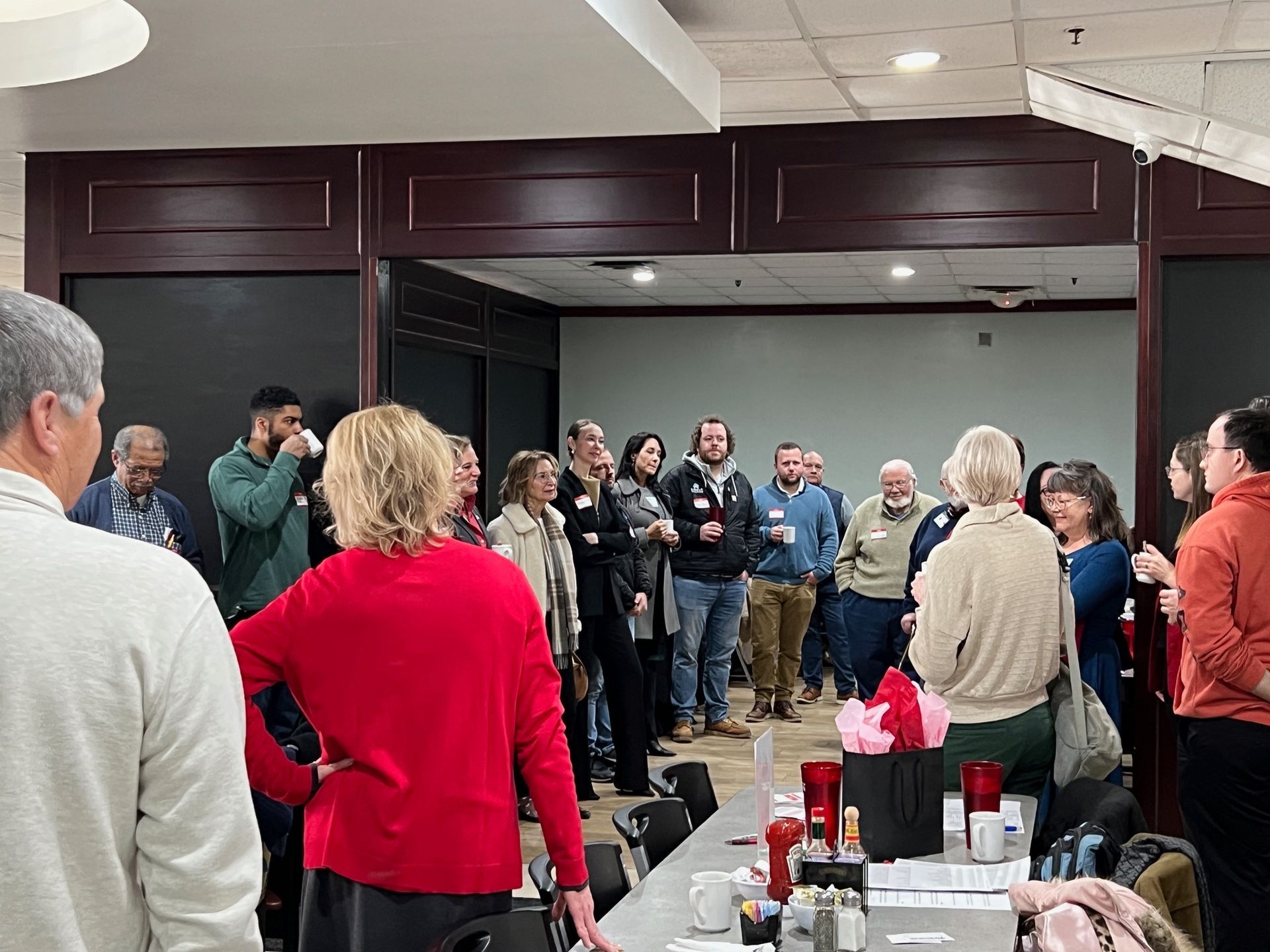 Group of people in a room, some standing, some at a table. Wooden paneling, red sweater, and casual attire are visible.