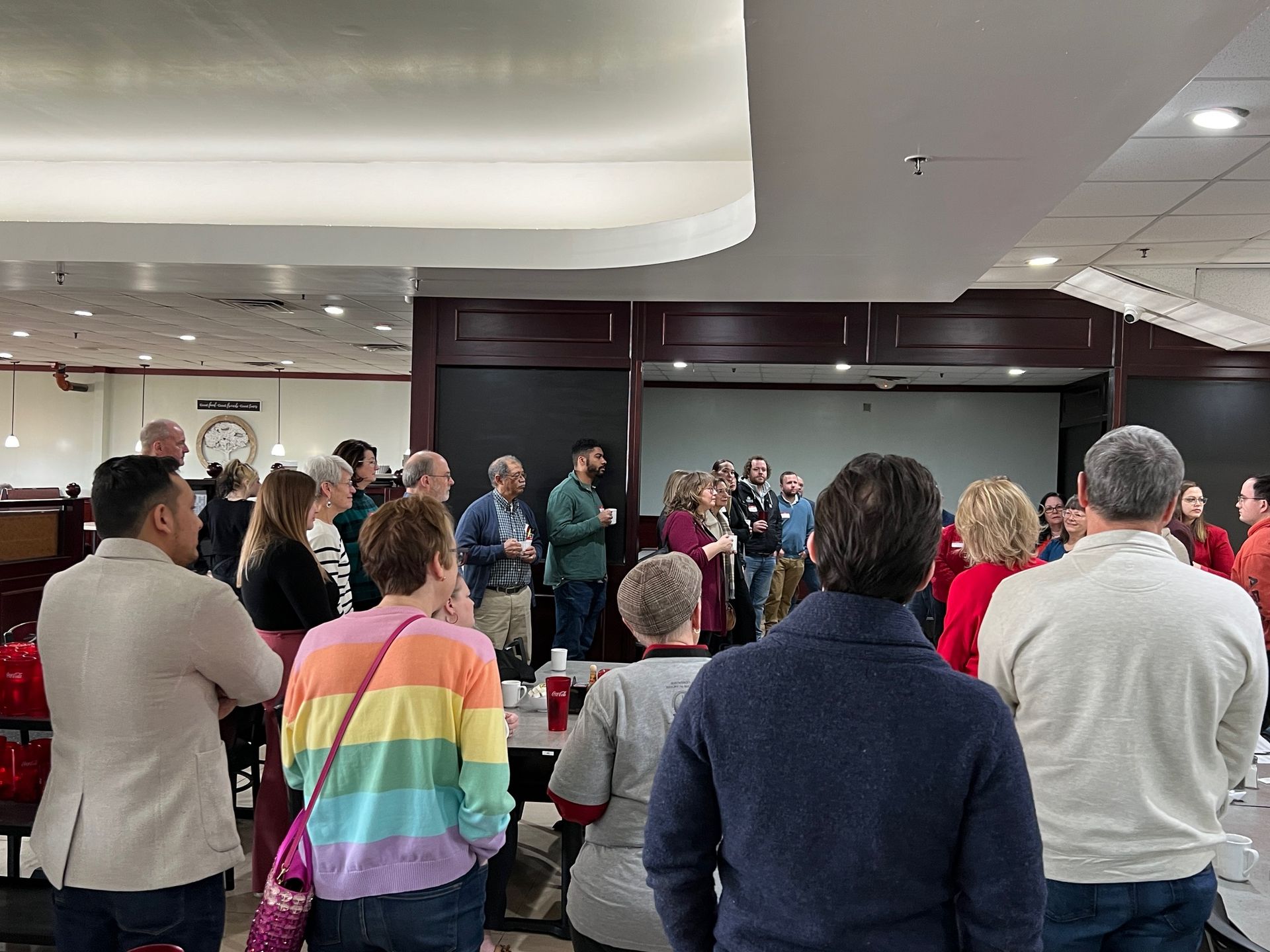 Group of people gathered indoors, listening to two people speak. Neutral setting with bright overhead lighting.