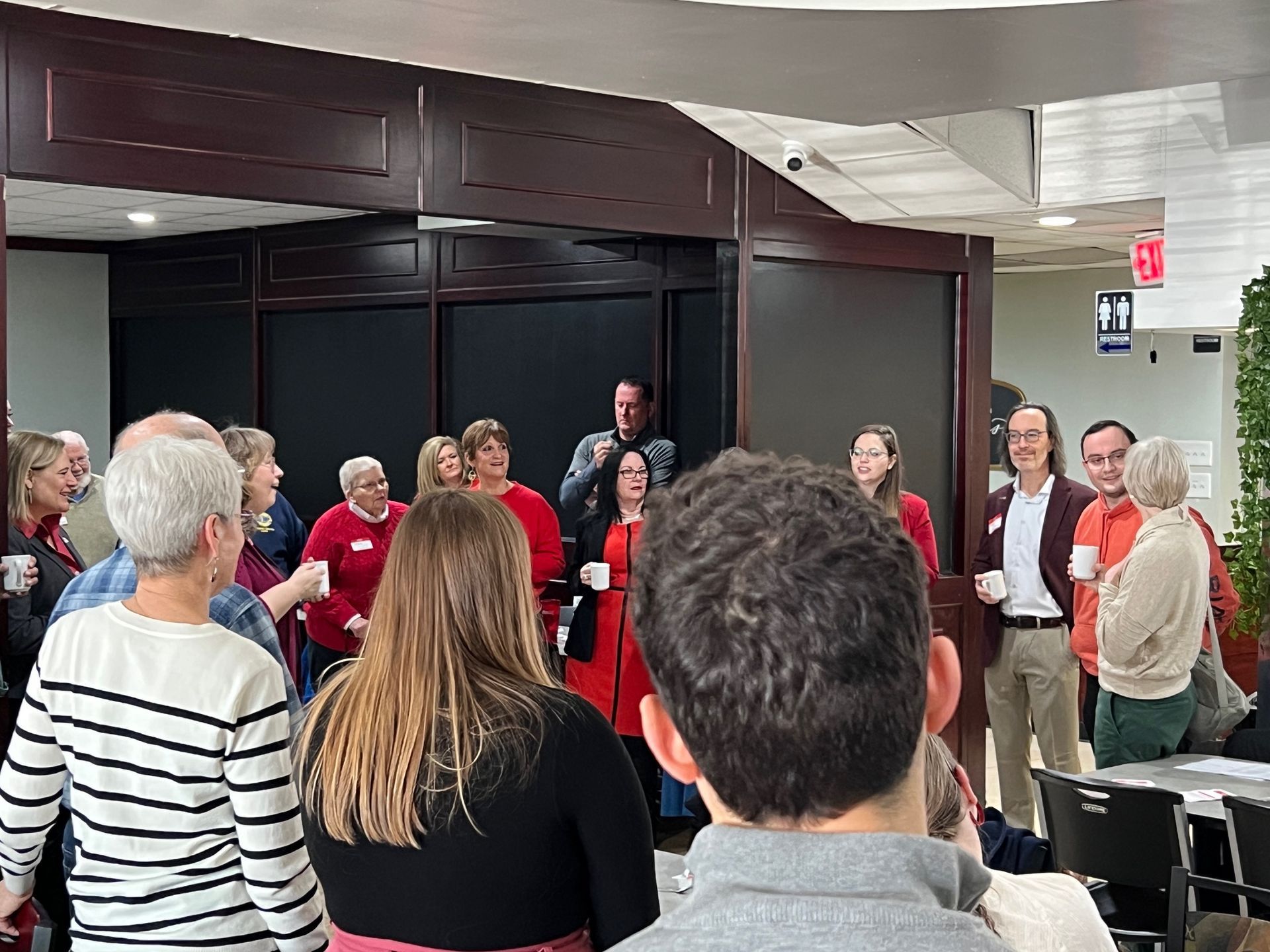 Group of people in a lobby, some holding mugs, gathered near a door. Many wear red shirts.