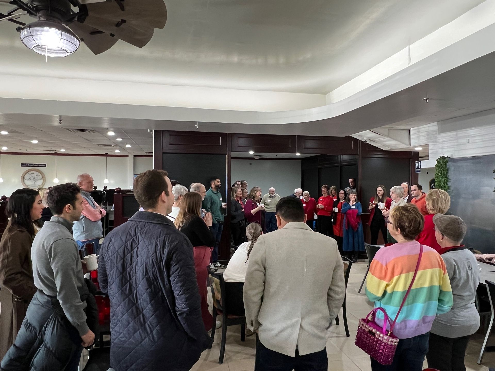 People gathered inside, some listening to a choir singing, near tables and a counter.