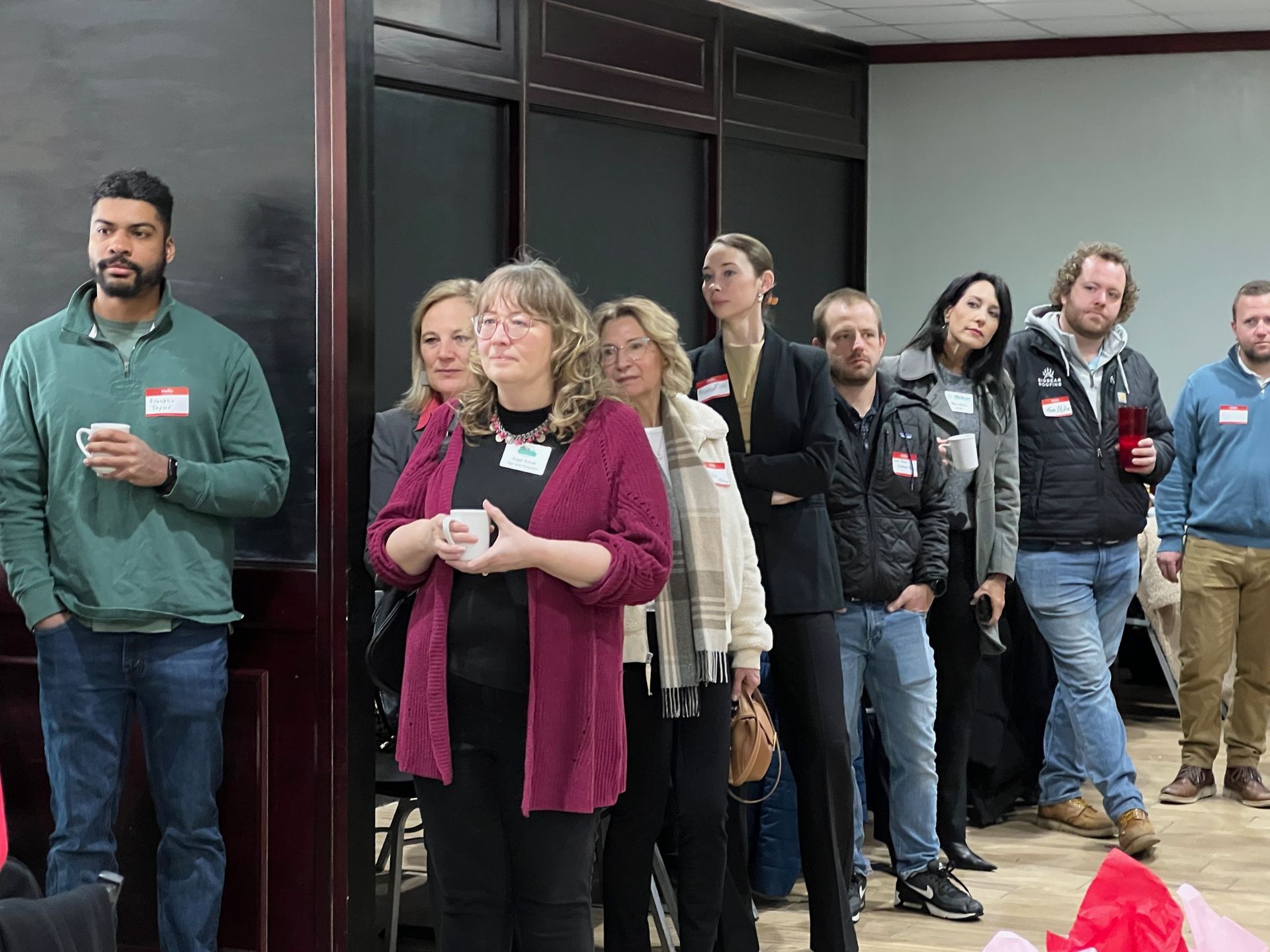 Group of people listening in a room. Some are holding drinks, wearing name tags.