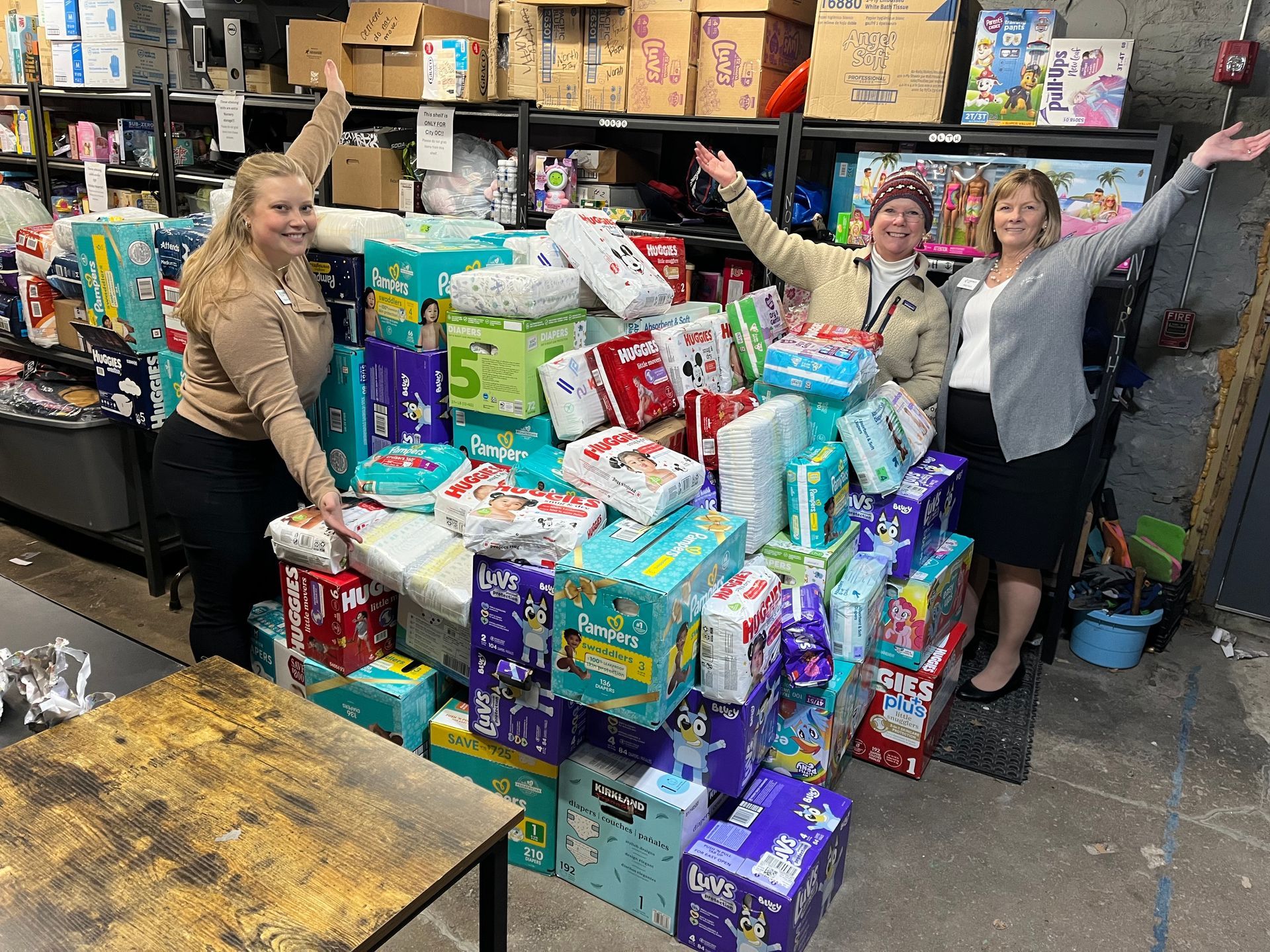 Three people stand with arms raised, surrounded by a large pile of diaper boxes in a storage room.