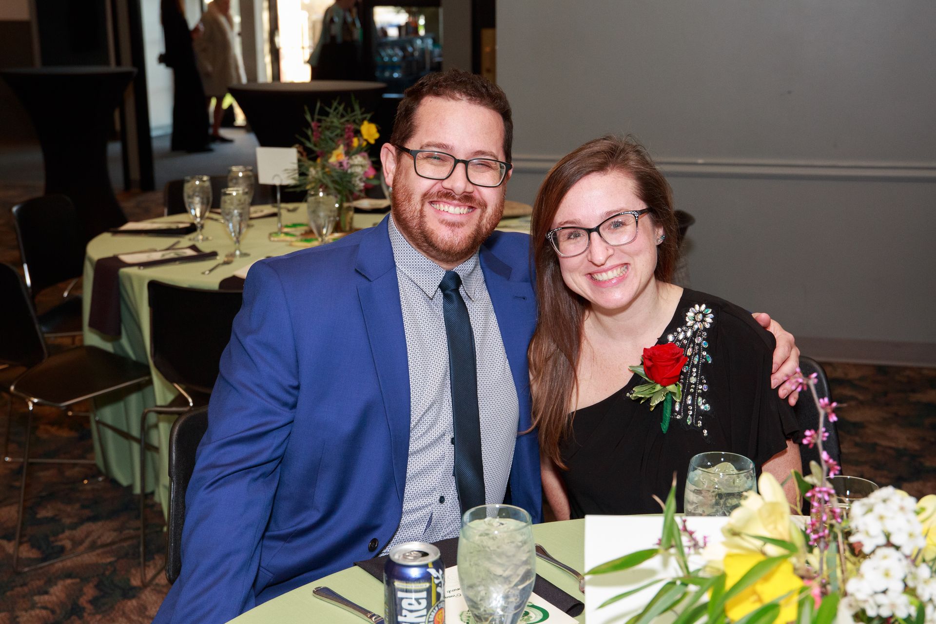 Man in blue suit and woman in black dress smile at a table with flowers.