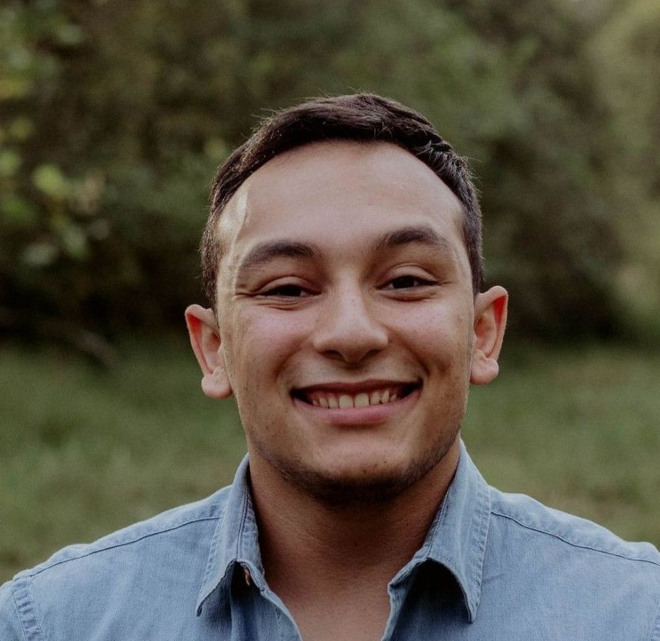 Man smiling outdoors, wearing a blue denim shirt, with a blurred natural background.