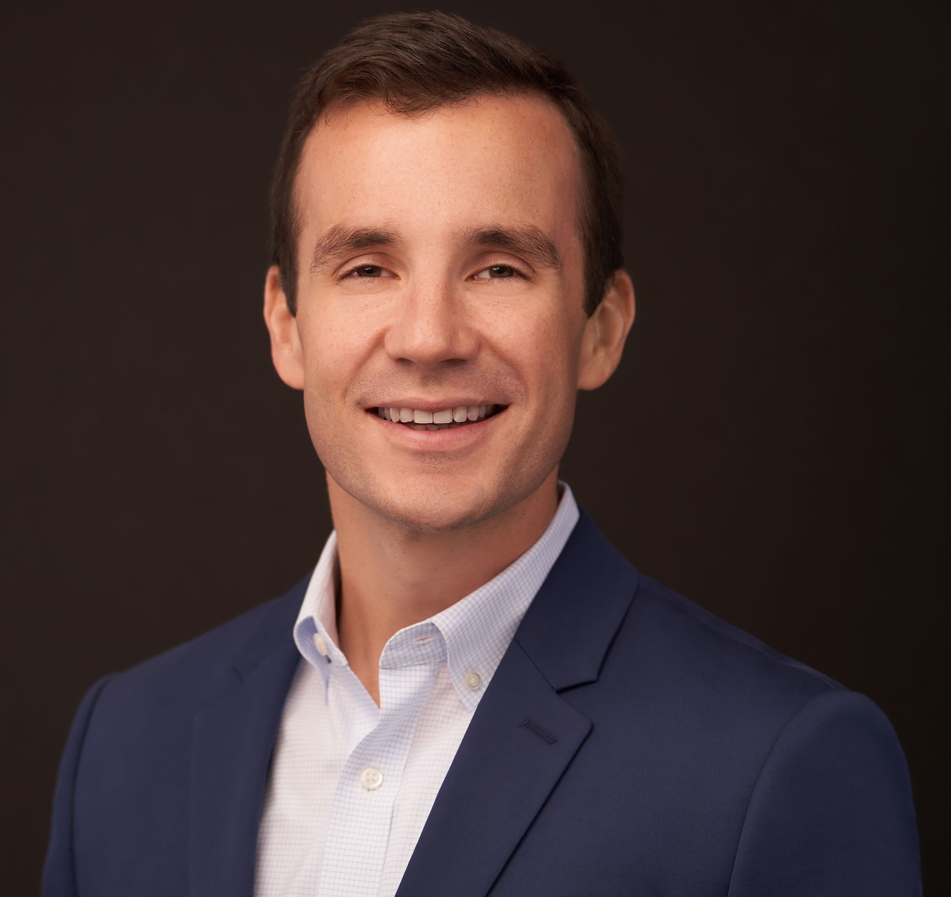 Man in a blue blazer and white shirt, smiling, against a dark background.