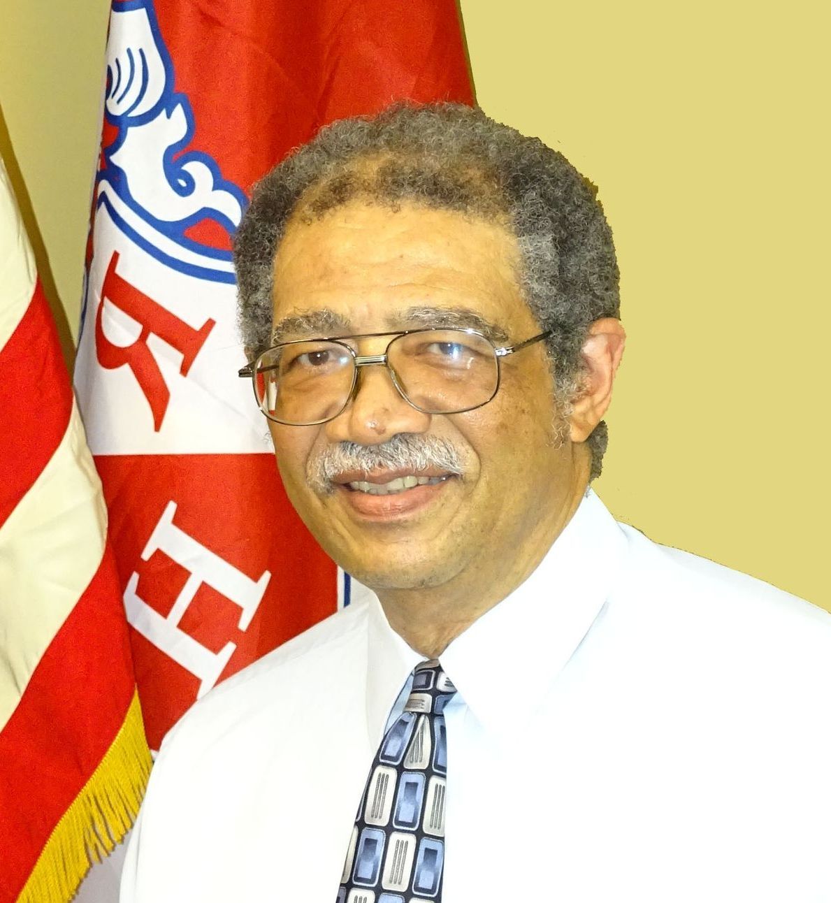 Man in glasses smiling, wearing a light blue tie and white shirt, flags in the background.