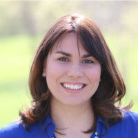 Woman with shoulder-length brown hair smiling broadly against a blurred outdoor background, wearing a blue shirt.