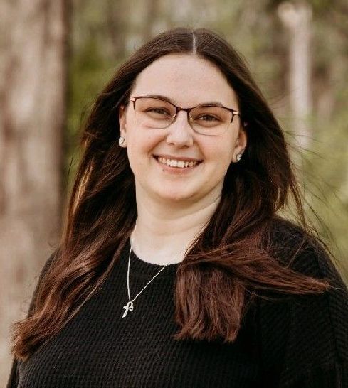 Woman with glasses smiles; wearing black sweater, silver cross necklace, trees in background.