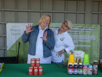 Two women at a booth with drinks, smiling, under a banner. Red cans stacked, colorful bottled drinks displayed.
