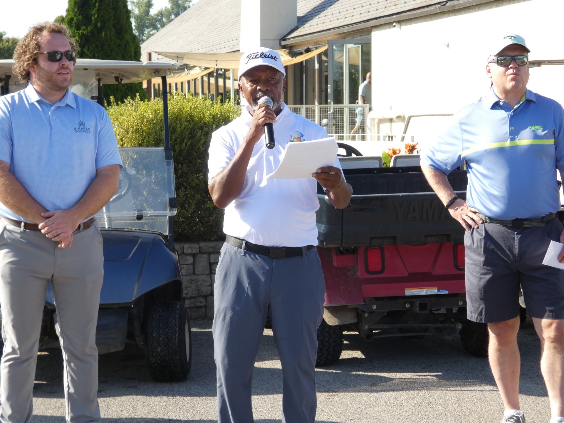 Three men at a golf outing. One speaks into a microphone, while the others stand nearby.