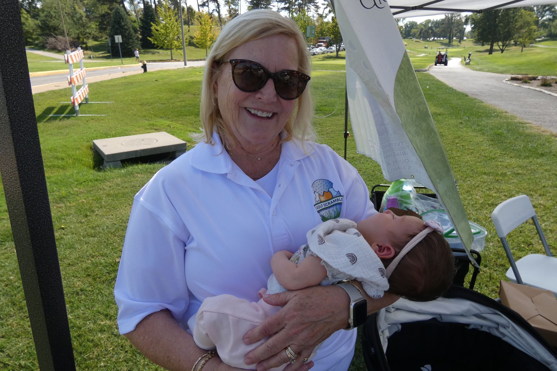 Woman in sunglasses smiles, holding baby outside. Green grass background.