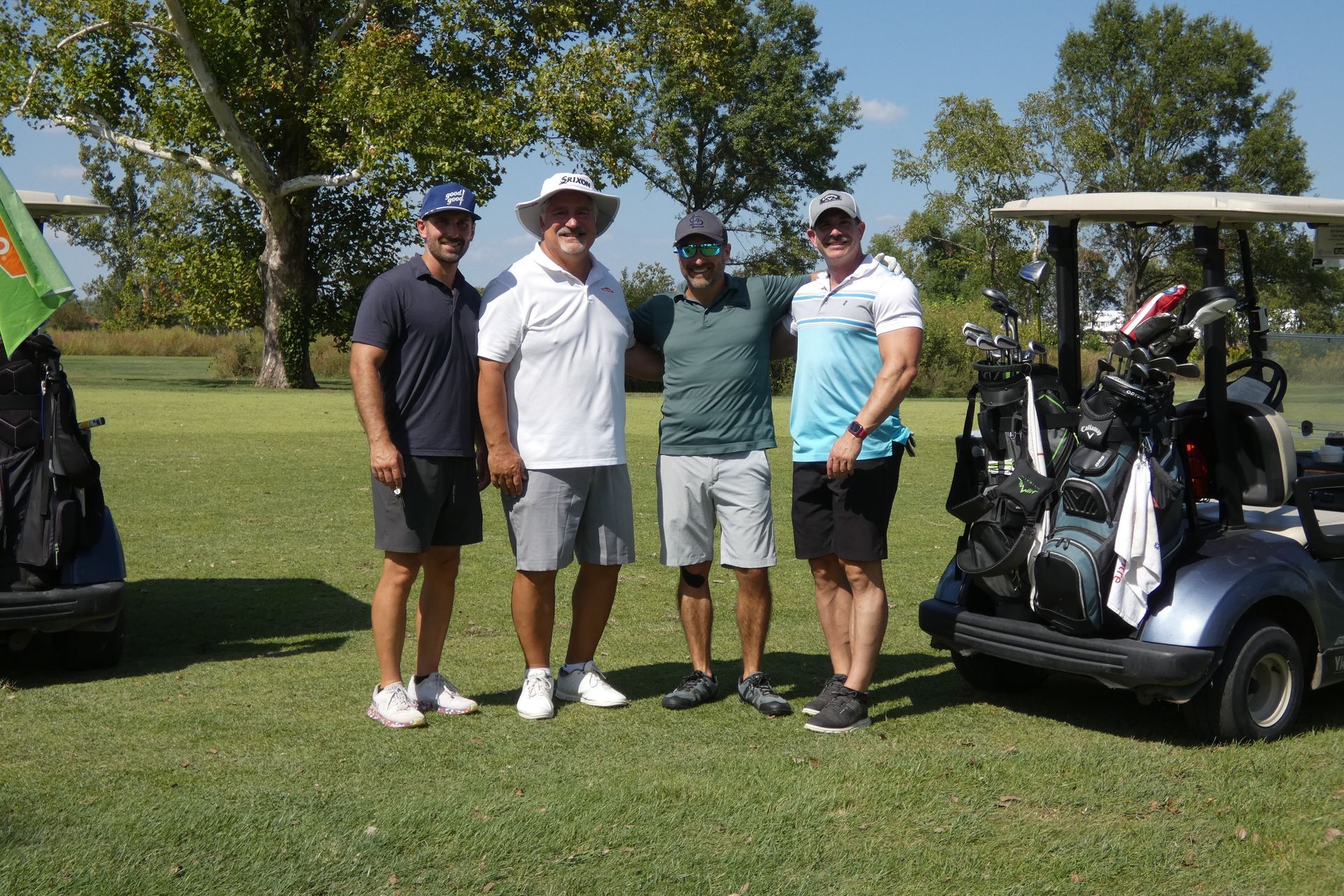 Four men pose on a golf course next to golf carts. Sunny day, green grass, casual attire.