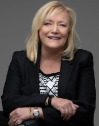 Woman with blonde hair wearing a black blazer and patterned shirt, smiling, arms crossed.