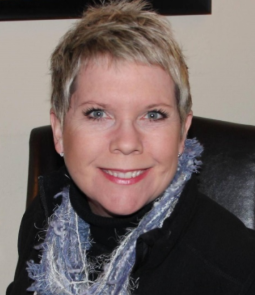 Woman with short blond hair, smiling. Wearing a black top and blue scarf. Sitting indoors.