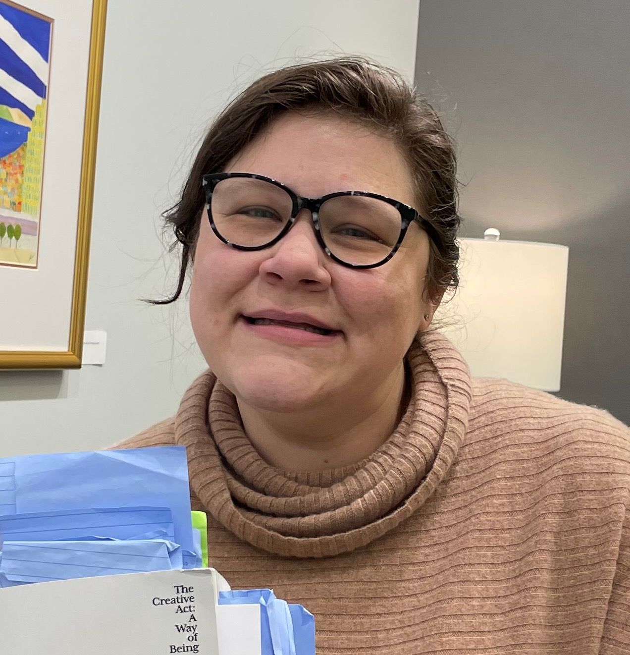 Woman wearing glasses and tan turtleneck smiles indoors near a lamp and painting, papers in foreground.