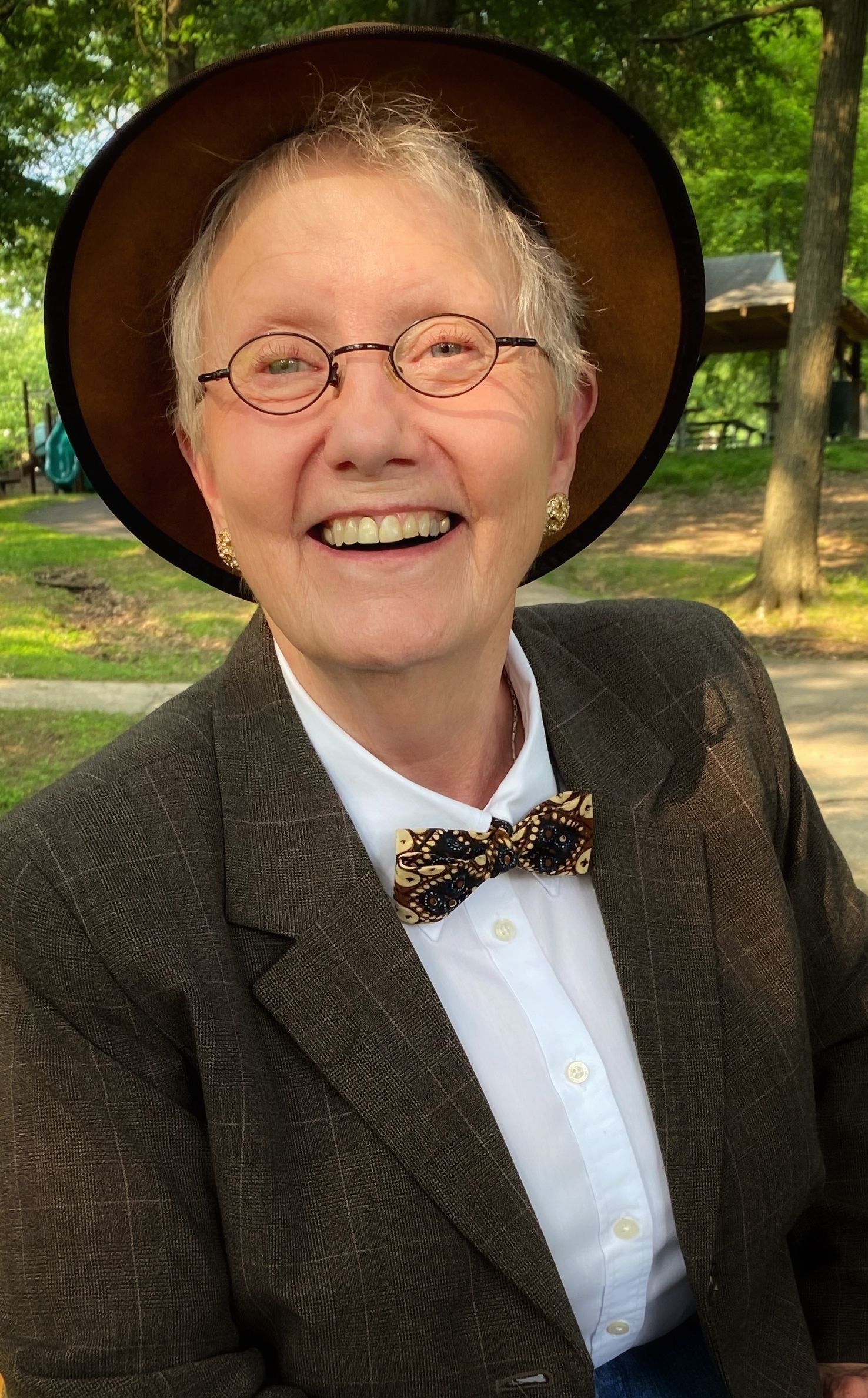 Woman smiling outdoors, wearing glasses, hat, tweed jacket, and bow tie.