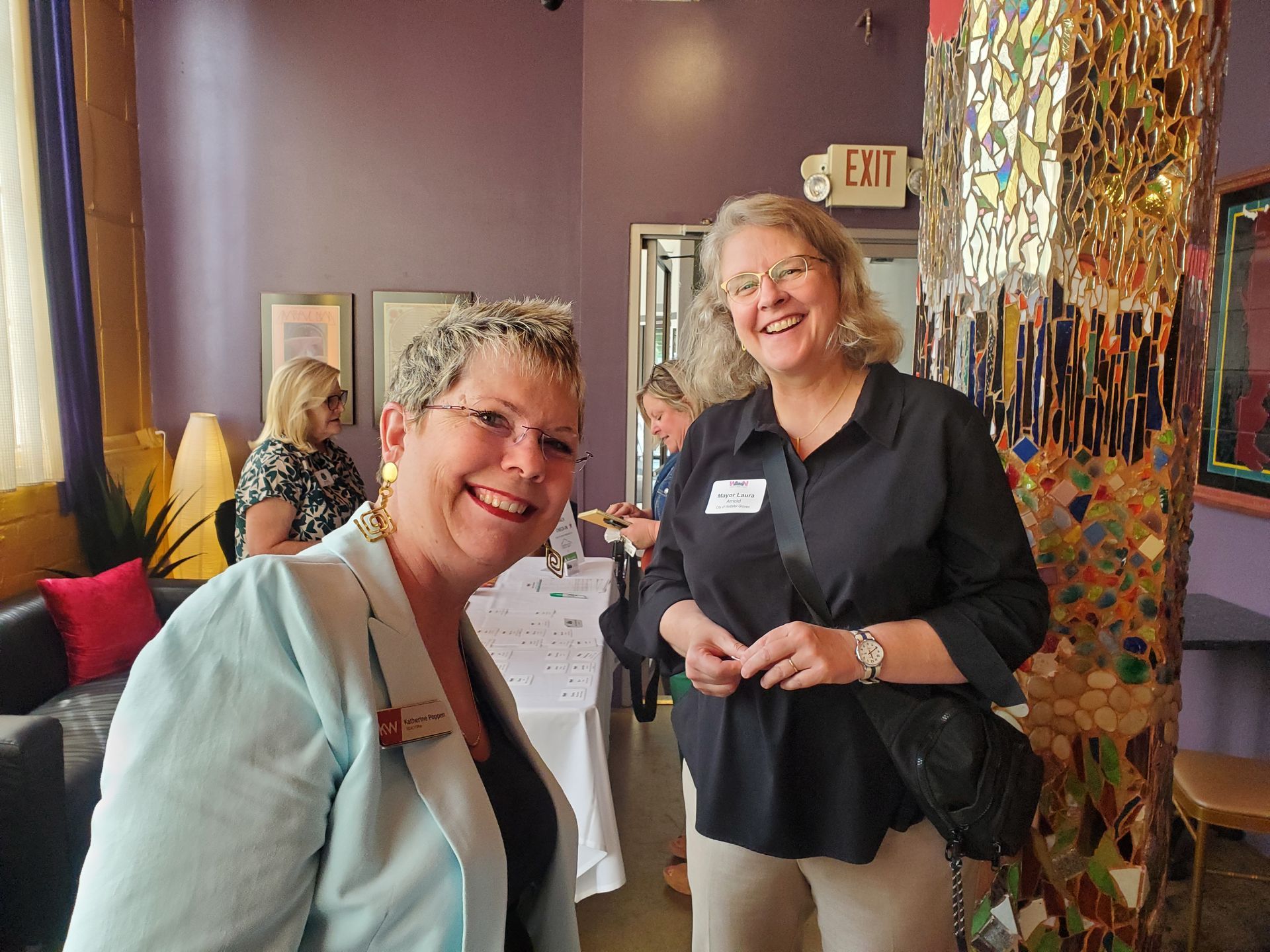 Two women smiling at an indoor event, one in a light blue jacket, the other in black. Purple wall, mosaic pillar.