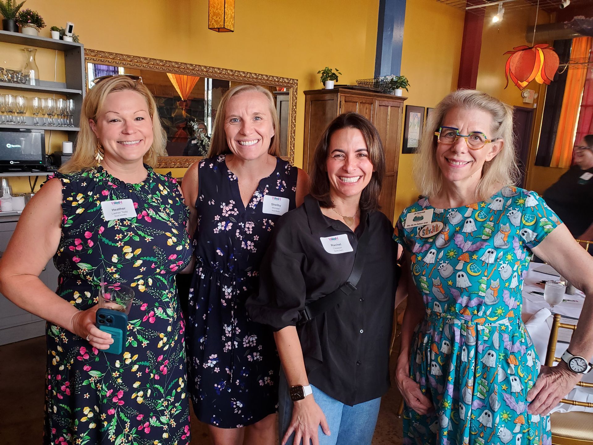 Four women smiling at the camera, inside a restaurant. They wear dresses and name tags.
