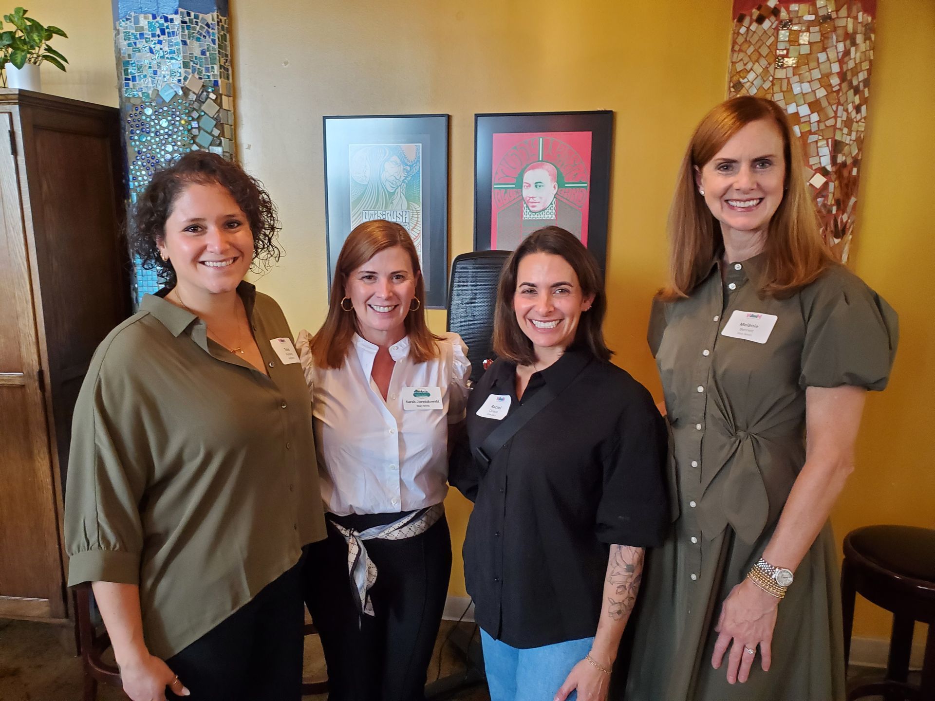 Four women smiling, posing inside a building. Two pictures on the wall behind them.