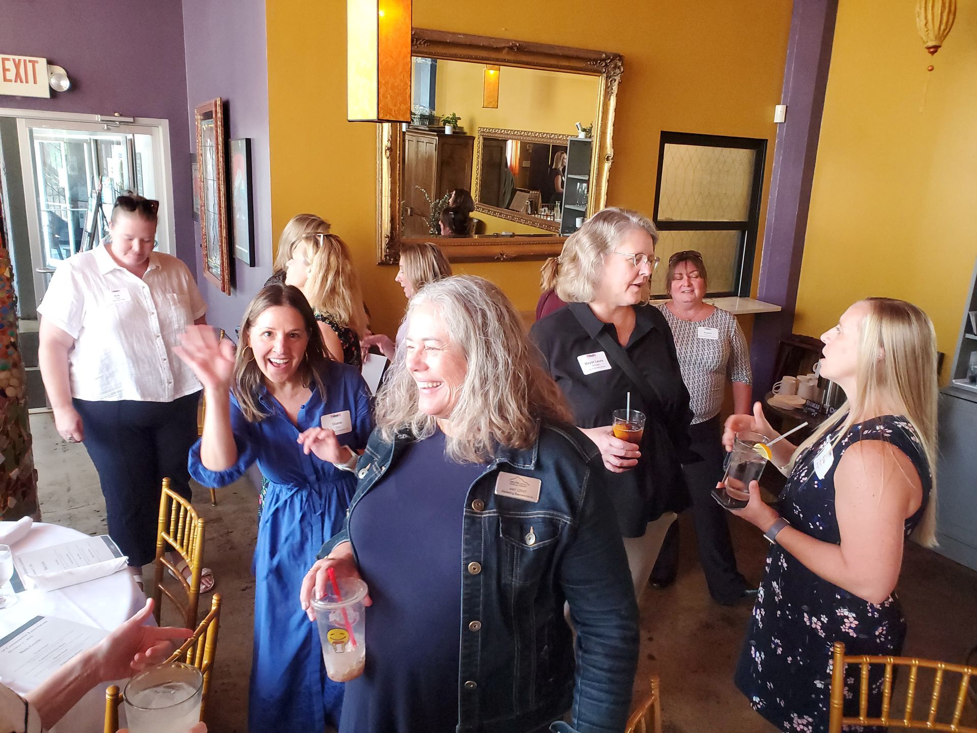 People in a restaurant setting. Some are chatting, waving, and holding drinks. Gold mirror and yellow walls.