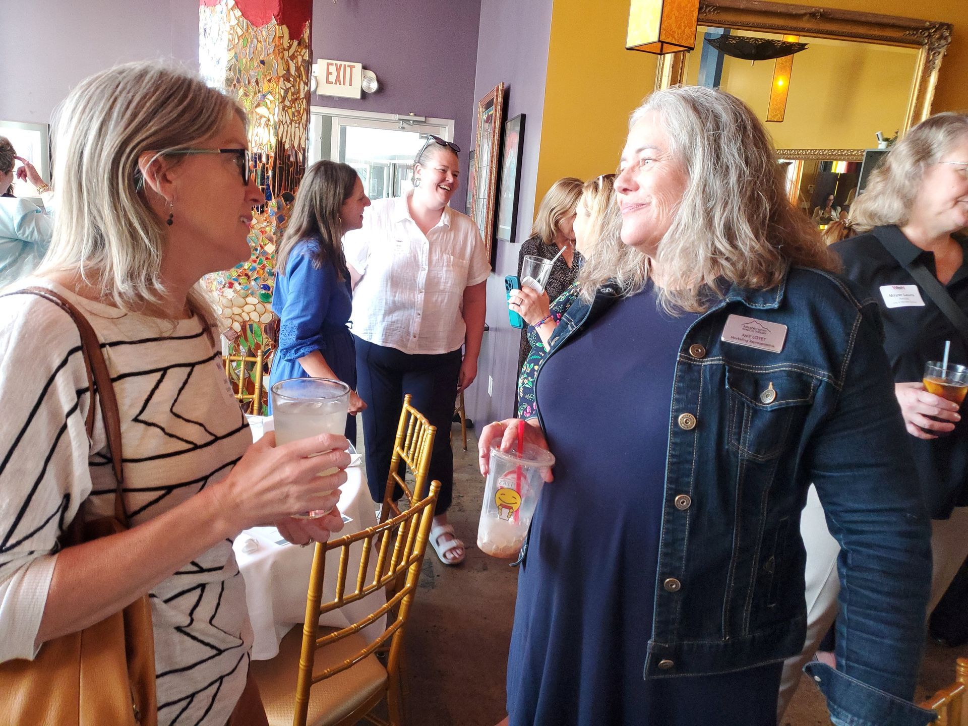 Two women conversing at an event, holding drinks. Other people and decorative interior in the background.