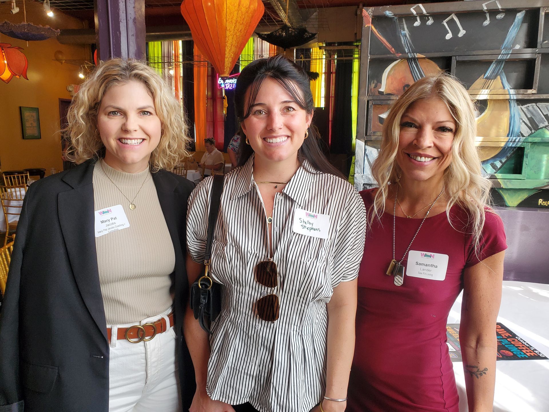 Three women smiling at the camera, inside a restaurant. One wears a blazer, another a zebra print top, the third a red dress.