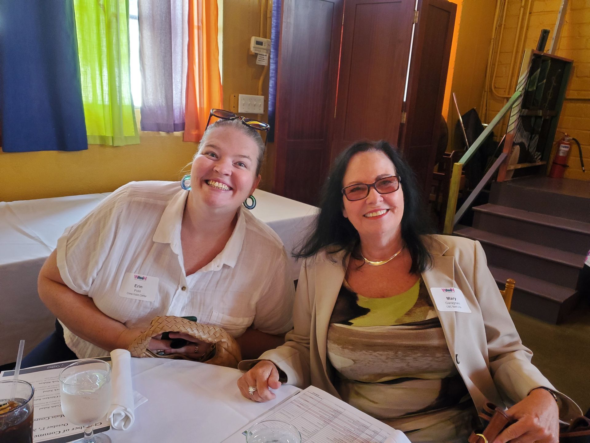 Two smiling women seated at a table. One wears a light shirt, the other a blazer. Indoor setting with colorful curtains.