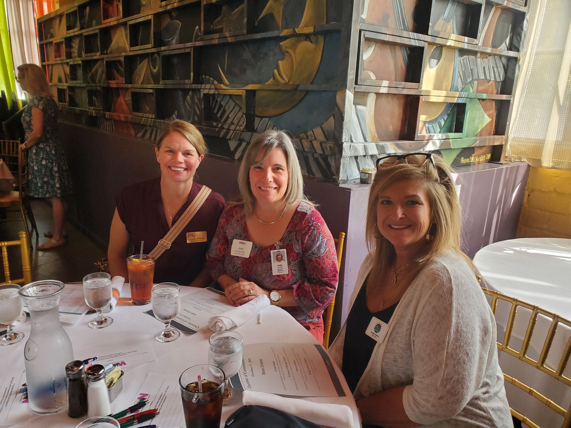 Three women at a restaurant table, smiling. They have name tags, drinks, and menus. Ornate wall in the background.