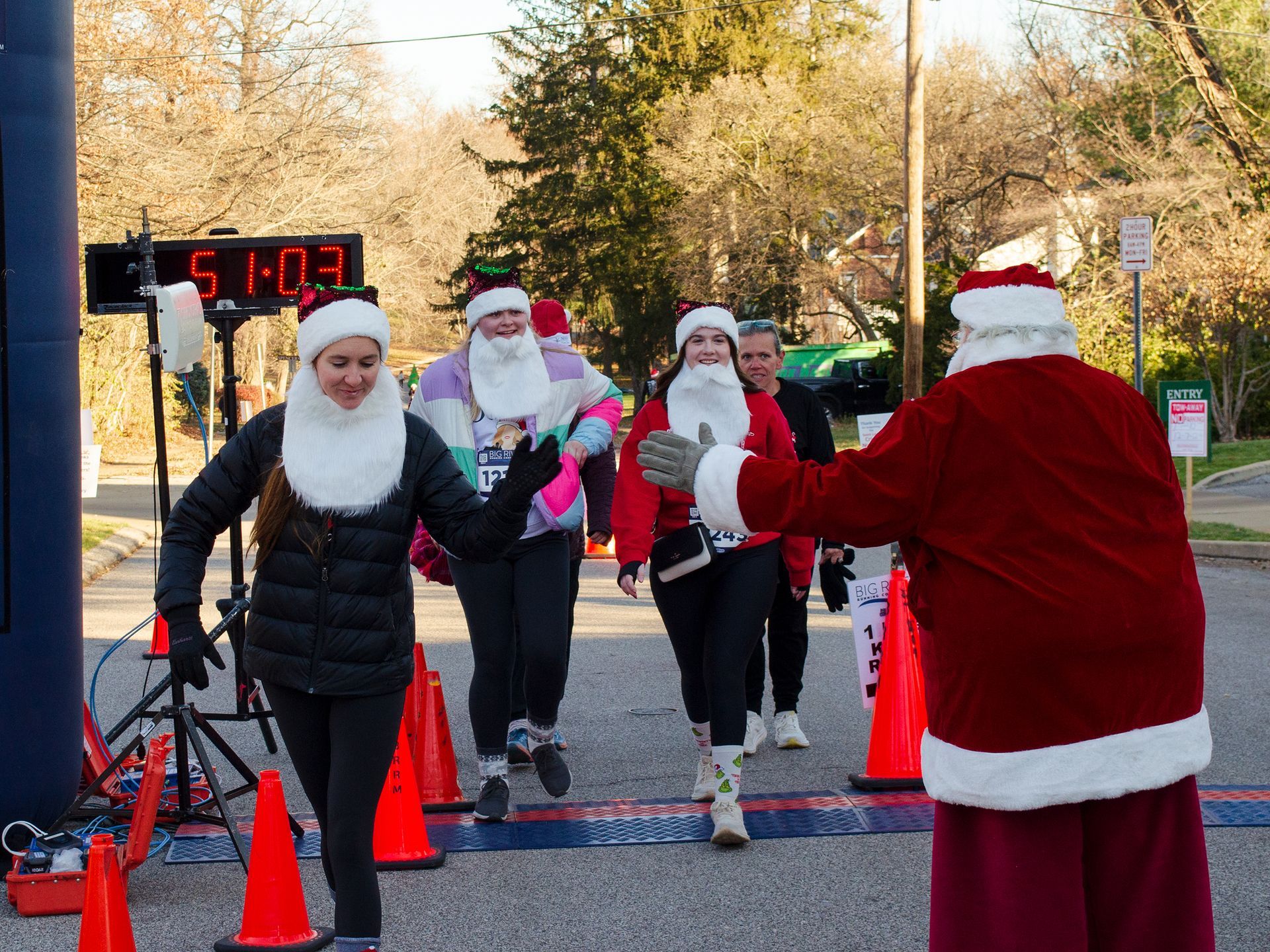 Runners in Santa hats and beards cross a finish line, greeted by Santa.