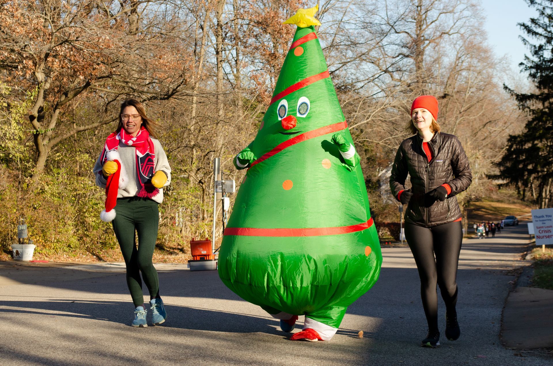 Two runners and a person in an inflatable Christmas tree costume on a road, trees in the background.