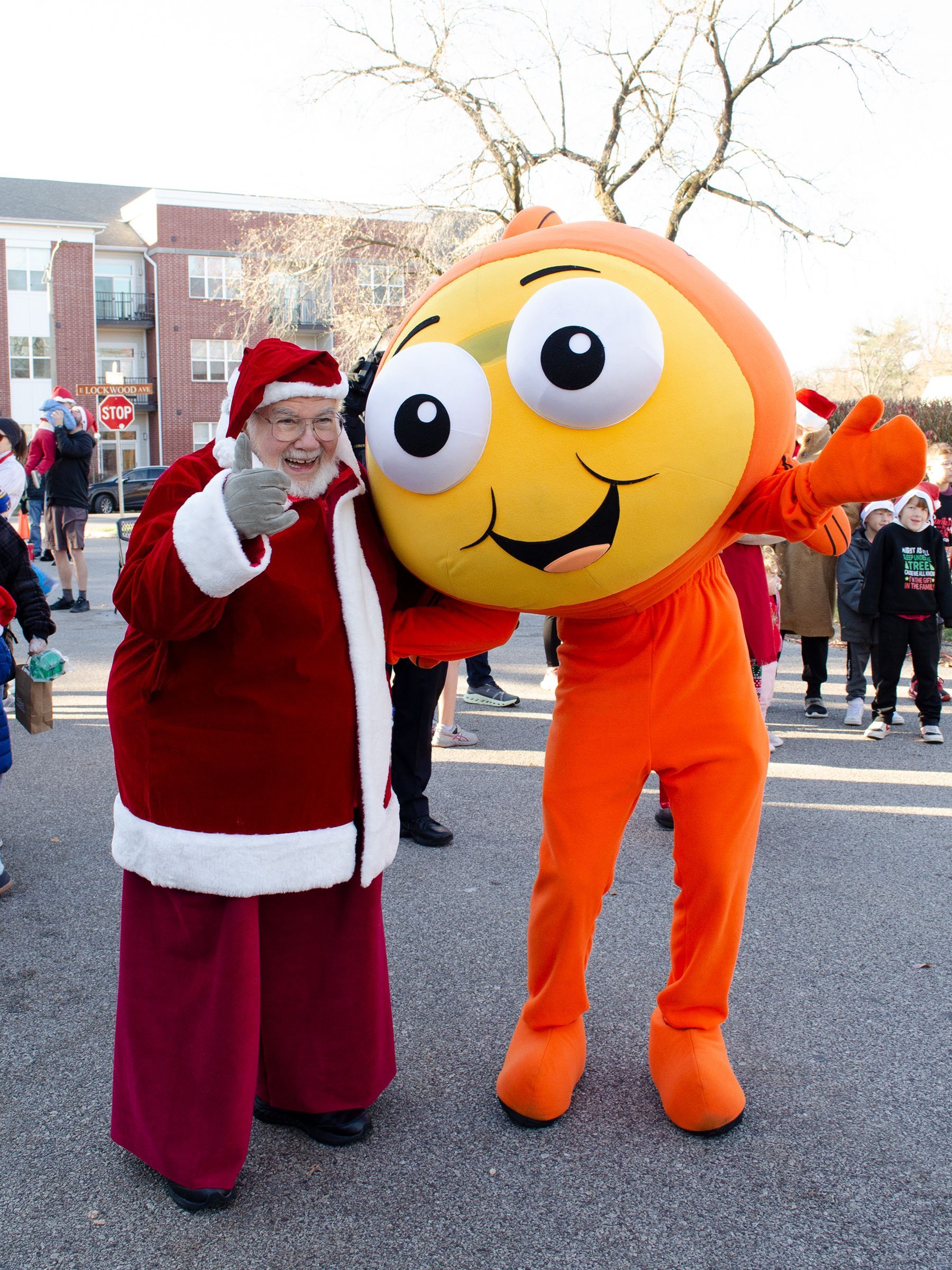 Santa Claus gives a thumbs-up next to a smiling orange mascot in an outdoor setting.