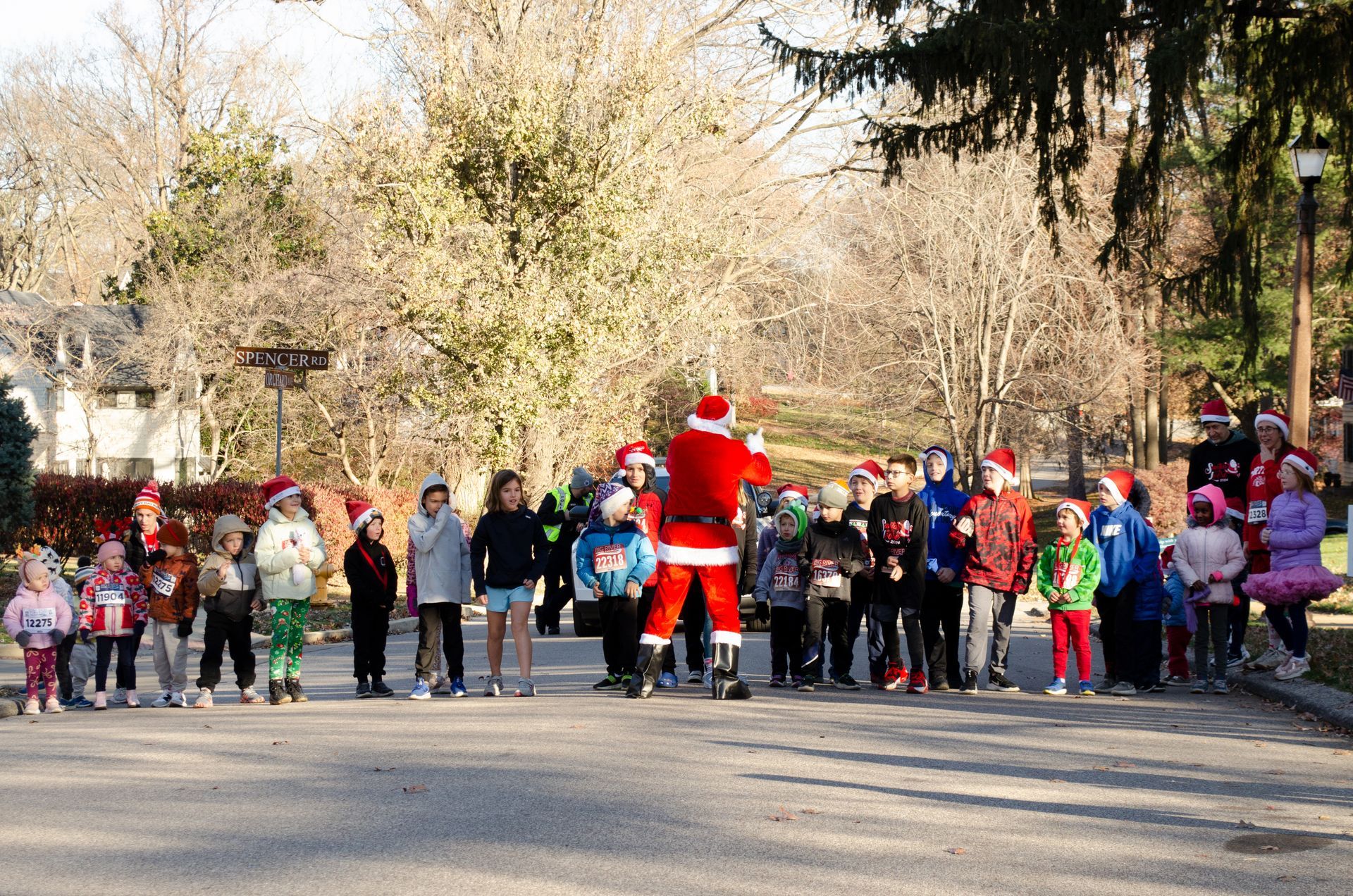 Santa Claus leads a group of children wearing Santa hats on a road lined with bare trees, preparing to run.