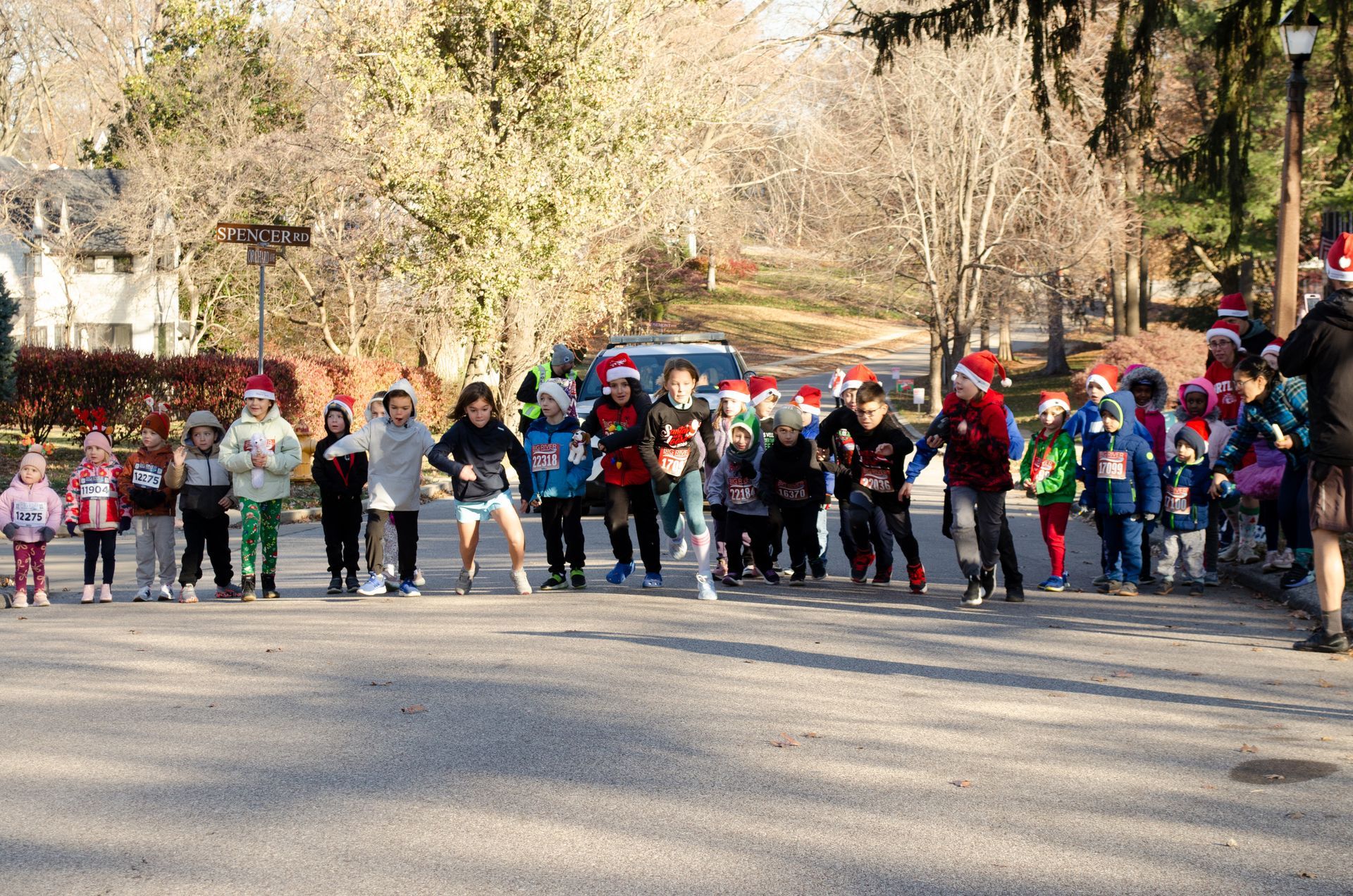 Children wearing Santa hats line up on a road, preparing for a race in a sunny outdoor setting.