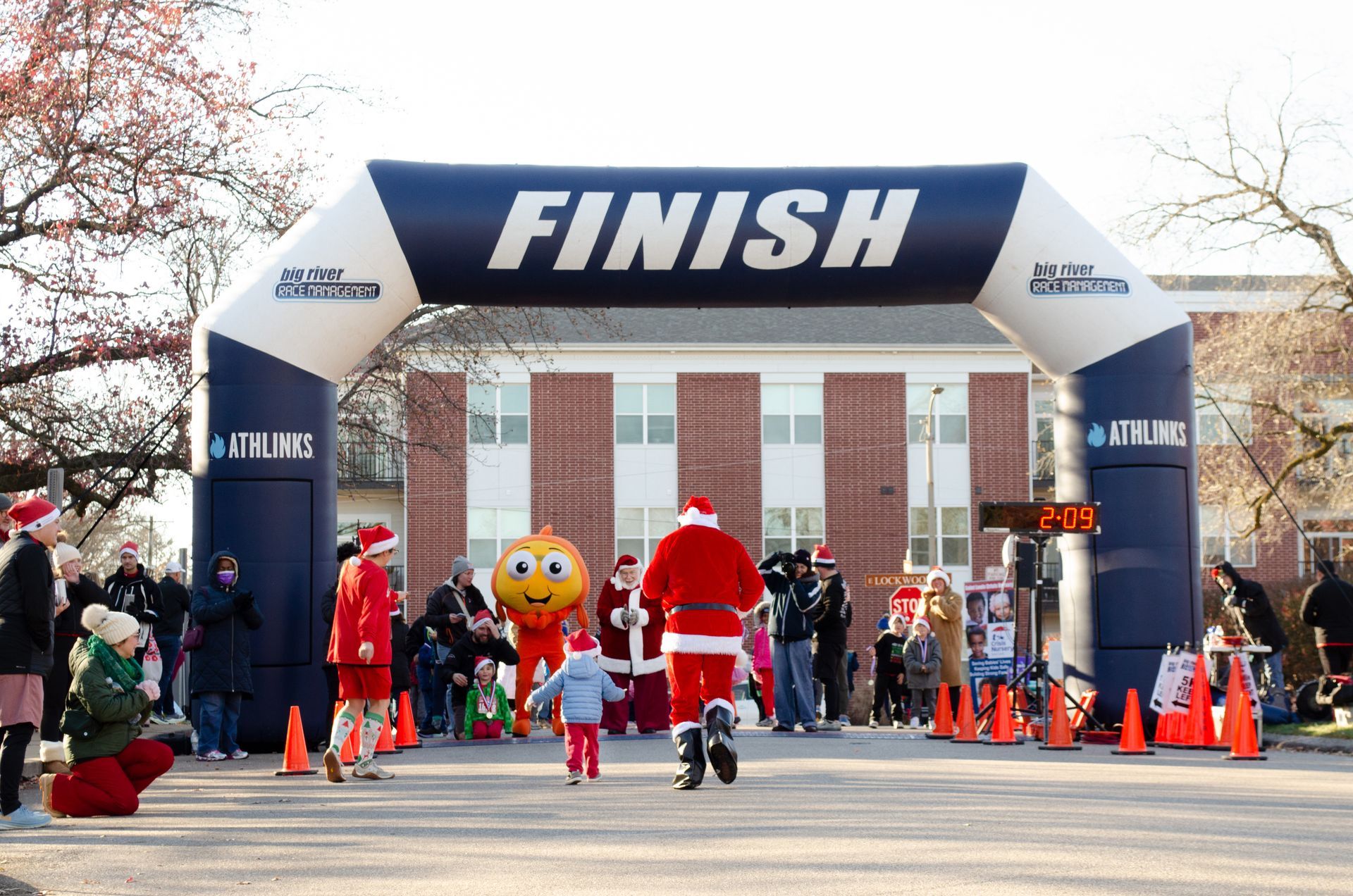 Runners in Santa suits approaching finish line at outdoor event.