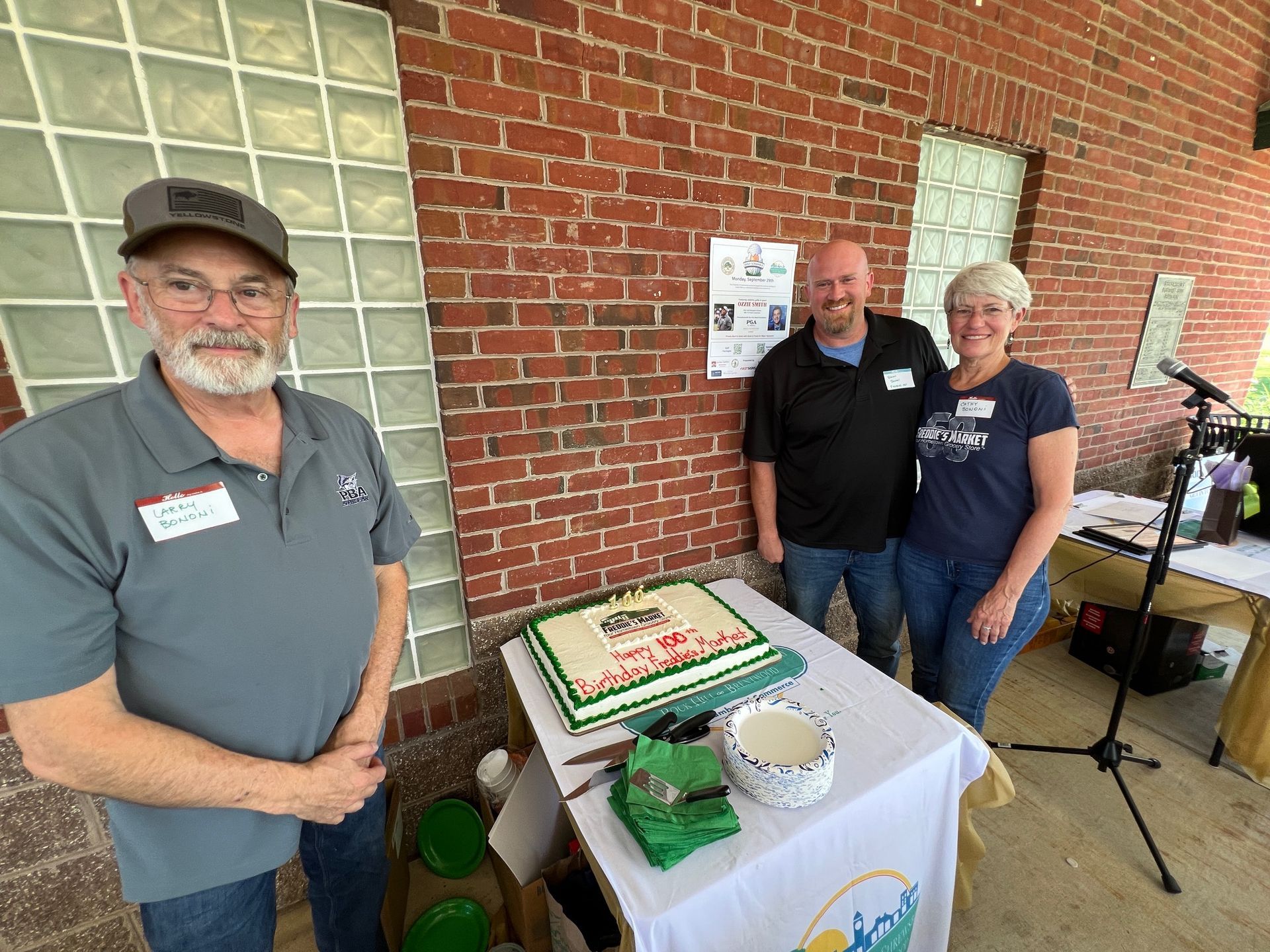 Three people pose by a decorated cake on a table; brick wall background.