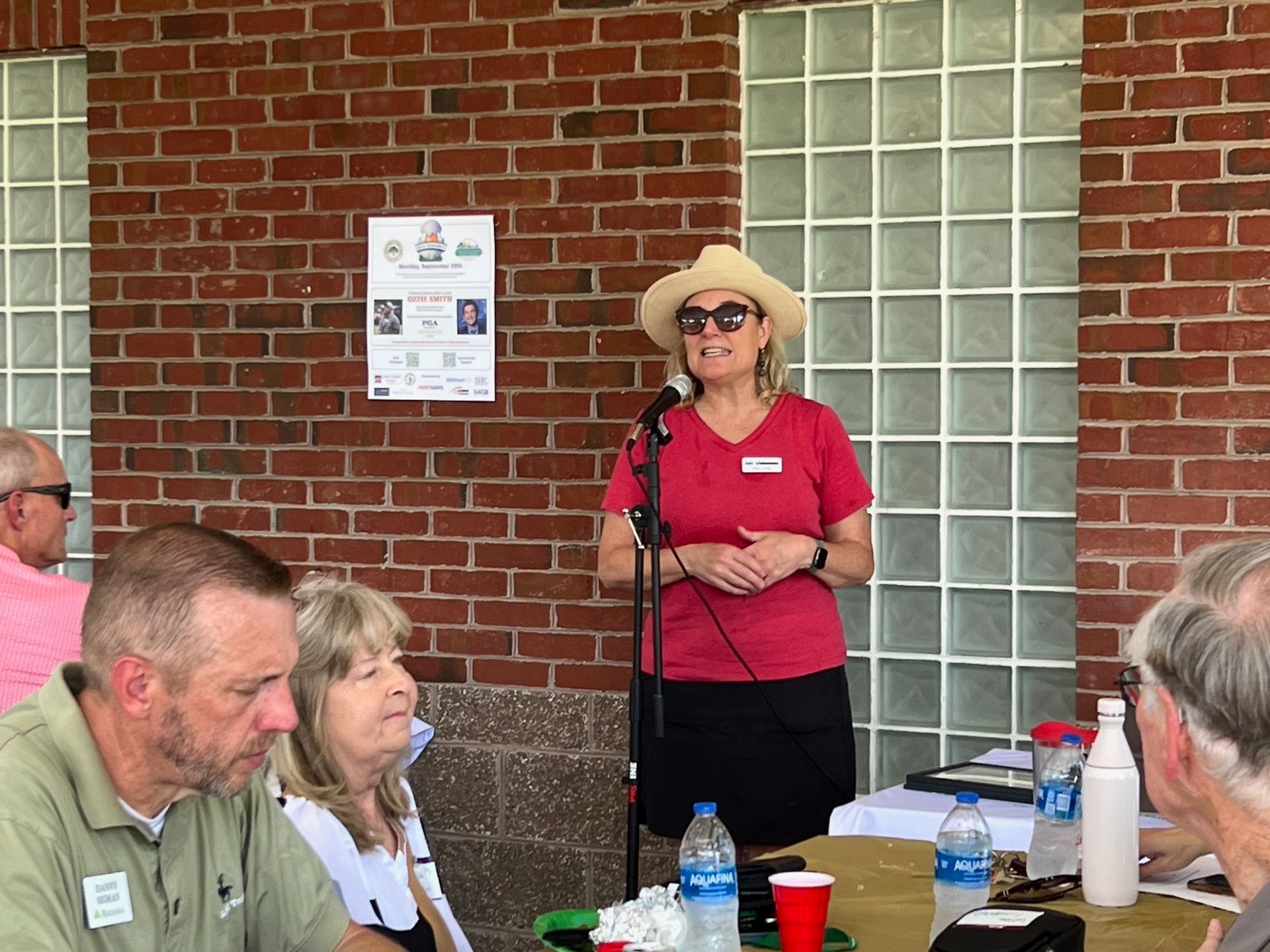 Woman in hat speaking at outdoor event, red shirt, facing an audience. Brick wall backdrop.
