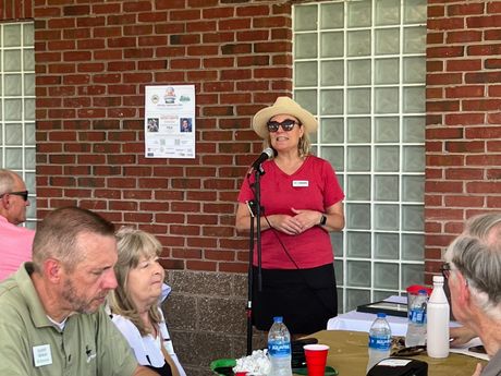 Woman in hat speaking at outdoor event, red shirt, facing an audience. Brick wall backdrop.