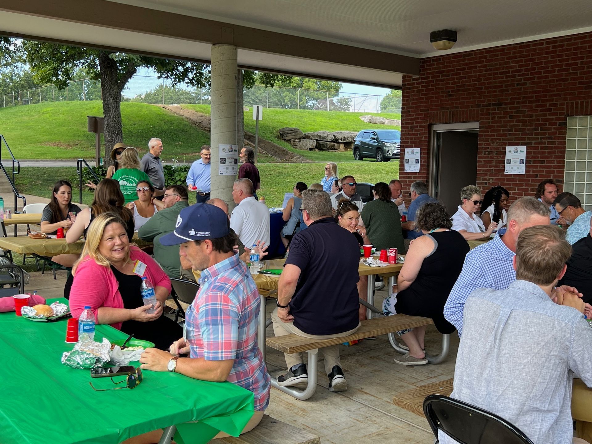 People gather at picnic tables under a pavilion for an outdoor event; food and drinks on tables.