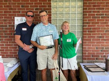 Three people stand near a brick wall; a man in a blue shirt holds a certificate as another in green speaks into a microphone.