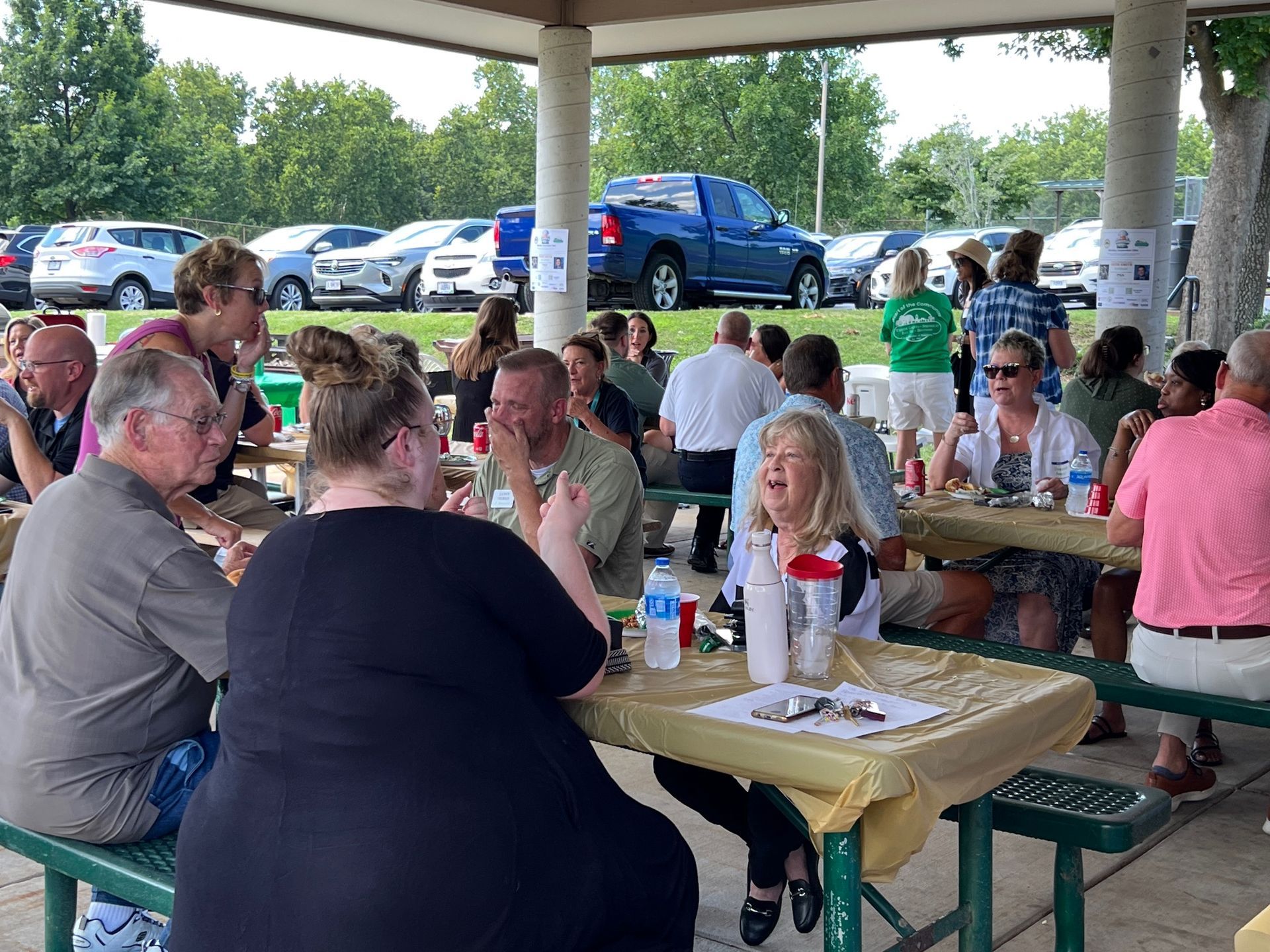 People sitting at picnic tables under a shelter, eating and socializing at an outdoor event. Cars and green space in background.